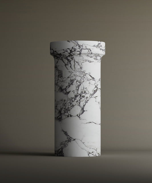 The TRAVERTABLE Calacatta Marble Pedestal Vanity Sink, featuring a tall cylindrical shape with a flared top and striking dark gray veining on a white marble-like base, stands against a plain brown background.