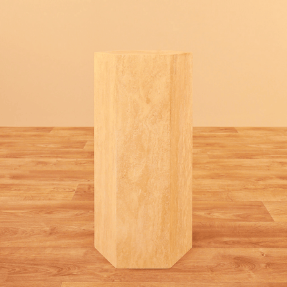 The Geometric Travertine Pedestal by Travertable, featuring a unique sculptural design and architectural appeal, stands tall like a hexagonal column on a matching wooden floor against a beige background.