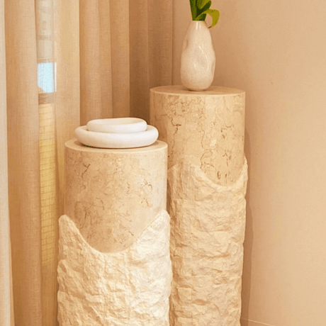 Two Rustic Charm Travertine Pedestals by Travertable stand against a curtain backdrop. A white vase with green leaves is on the taller one, while stacked white rings adorn the shorter, crafting a minimalist and modern display of sculptural columns.