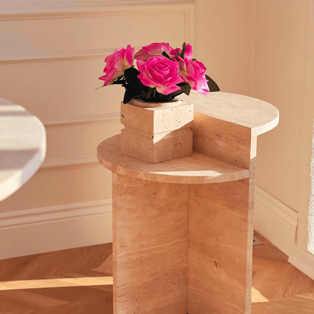 A bouquet of vibrant pink roses in a beige stone vase rests gracefully on the Travertable Contemporary Travertine Side Table, stylishly versatile for modern living. The scene is bathed in warm sunlight, casting soft shadows on the wooden floor and white paneled walls, creating a serene interior.