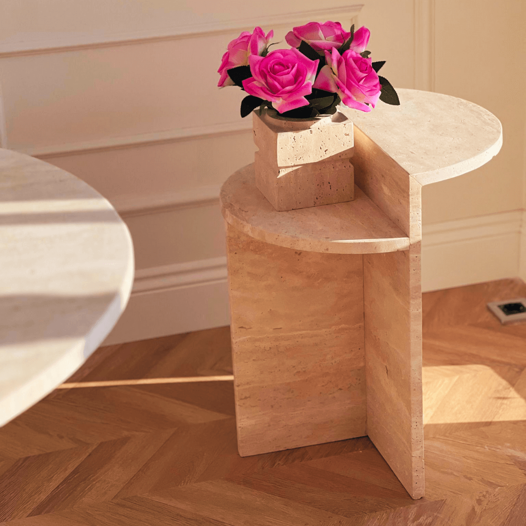 The sleek Contemporary Travertine Side Table by Travertable, featuring a unique geometric design, complements a square vase of vibrant pink roses. This sophisticated arrangement rests on a wooden herringbone-patterned floor, beautifully illuminated by soft natural lighting.