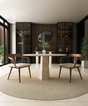 A modern dining room features the TRAVERTABLE Oval Travertine Dining Table with Rockcone Center Base, two wooden chairs, a circular rug, built-in glass cabinets, a round mirror, and decorative items on a wood-paneled wall.