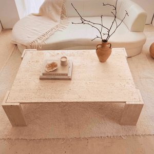 A minimalist living room showcases a white sofa and a Modern Rustic Travertine Coffee Table by Travertable, adorned with a vase of twigs, sculpture, candle, and stacked books. A textured rug and blanket over the sofa highlight the natural beauty of this expertly crafted cozy setting.