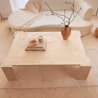 A minimalist living room showcases a white sofa and a Modern Rustic Travertine Coffee Table by Travertable, adorned with a vase of twigs, sculpture, candle, and stacked books. A textured rug and blanket over the sofa highlight the natural beauty of this expertly crafted cozy setting.