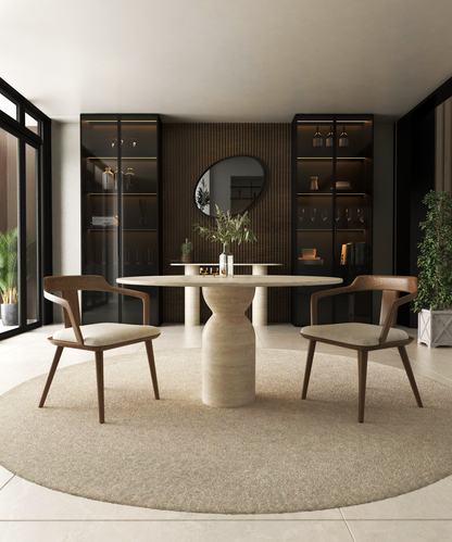 A modern dining room features the TRAVERTABLE Oval Travertine Dining Table with a sculptural round center base, two wooden chairs, a circular rug, built-in shelves, a large round mirror, and vases on the back wall illuminated by natural light.