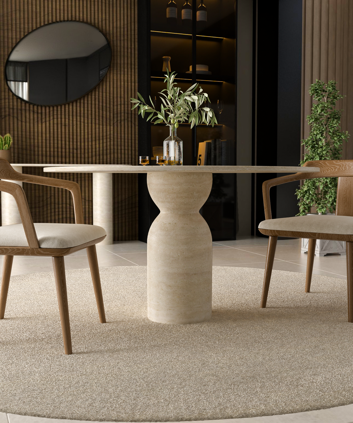 The TRAVERTABLE Oval Travertine Dining Table with a sculptural round center base stands on a beige rug between two wooden chairs. A glass bottle with branches and two glasses sit on the table, set against a wall mirror, plants, and a wooden accent wall.
