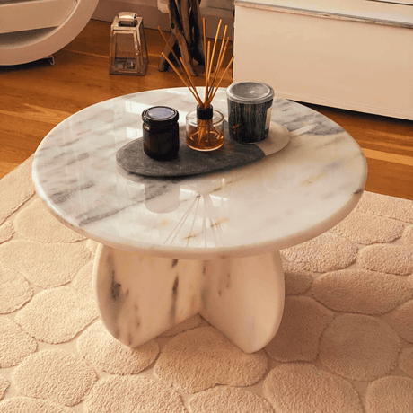 The Travertable Stone Essence Travertine Coffee Table, with its monolithic modern design, rests on a textured beige rug. Adorned with two dark candles and a reed diffuser set on a dark tray, the room highlights natural beauty with wooden flooring and minimal decor.