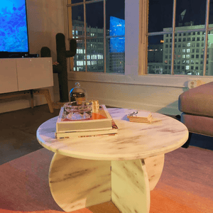 A cozy living room showcases a timeless design with a Travertable Stone Essence Travertine Coffee Table, styled with books and decor. Large windows display a vibrant cityscape, complemented by a cactus-shaped decor piece and TV, adding charm to the scene.