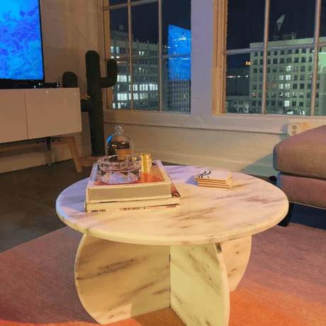 A cozy living room showcases a timeless design with a Travertable Stone Essence Travertine Coffee Table, styled with books and decor. Large windows display a vibrant cityscape, complemented by a cactus-shaped decor piece and TV, adding charm to the scene.