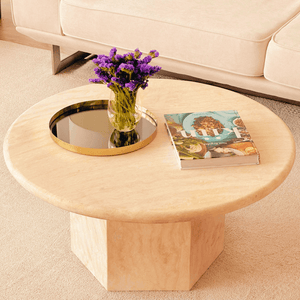 The Desert Serenity Travertine Coffee Table by Travertable, made from earthy stone with a honed finish, showcases a timeless design. It features a glass vase with purple flowers, a black and gold tray, and a colorful magazine on a light carpet in front of the cream sofa.