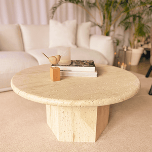 A Desert Serenity Travertine Coffee Table by Travertable, adorned with a small wooden bird sculpture and stacked books, graces the room. A white sofa and potted plants in the background enhance the timeless design, creating a cozy, modern living room atmosphere.