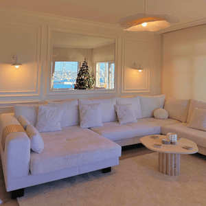 A cozy living room showcases a large beige sectional and a Timeless Stone Travertine Coffee Table by Travertable. A window reflects a decorated Christmas tree and marina view, while soft lighting from wall sconces enhances the warm ambiance.