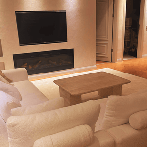 A cozy living room with a plush white sofa facing a flat-screen TV above a modern fireplace features Travertables Limestone Luxury Coffee Table, showcasing timeless natural beauty and sculptural design. Warm lighting and a partially open door reveal an elegant connection to another room.