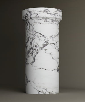 The TRAVERTABLE Calacatta Marble Pedestal Vanity Sink features a tall, cylindrical design with a wider top and elegant gray veining. The plain backdrop accentuates its timeless, sophisticated look.