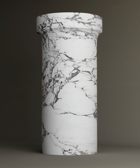The TRAVERTABLE Calacatta Marble Pedestal Vanity Sink features a tall, cylindrical design with a wider top and elegant gray veining. The plain backdrop accentuates its timeless, sophisticated look.