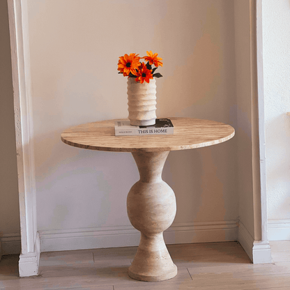 A minimalist setting features Travertables The Hourglass Round Travertine Dining Table against a neutral-hued wall, creating a serene vibe. It holds a white vase with bright orange flowers and two books, including This is Home.