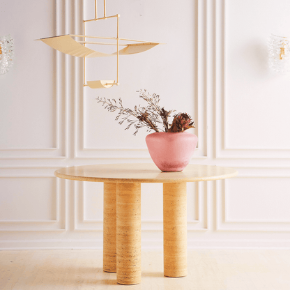 The Trio Round Travertine Dining Table by Travertable with dual cylindrical legs is set against white paneled walls. A pink vase with dried flowers sits atop, while a modern geometric pendant light illuminates the light wood floor below.