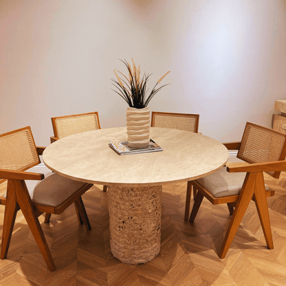 The Rustic Round Travertine Dining Table by Travertable features a textured pedestal stone base on wooden parquet. Four wooden chairs with woven backs and cushions surround the customizable tabletop, decorated with a plant and magazines.