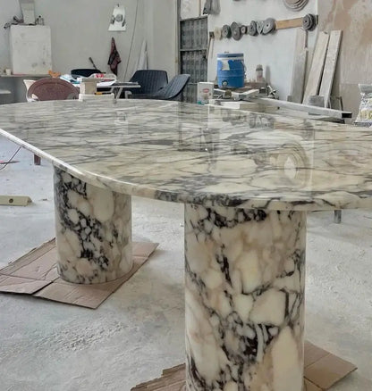 The Arcadia Dining Table by Travertable, with its polished marble rectangular top and two cylindrical pedestals in a black and white pattern, stands proudly in the workshop. Customize your table to suit any space with tools and chairs visible in the background.