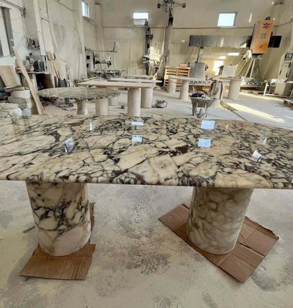 In a workshop, an Arcadia Dining Table by Travertable features a glossy, patterned marble surface on two cylindrical bases. It invites customization amid stonework materials and tools, alongside partially assembled travertine dining pieces.