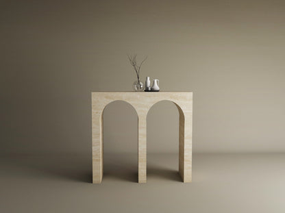 The TRAVERTABLE Travertine Console Table with Two Arches—a solid stone, customizable piece—stands against a taupe backdrop, topped with a clear glass vase of bare branches and two small white ceramic vases.
