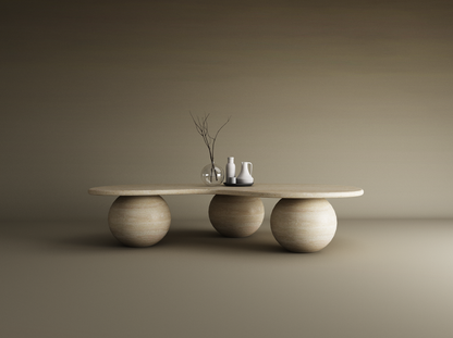 The TRAVERTABLE Wavy Travertine Coffee Table features an irregular oval top and three stone ball bases. Styled with a glass vase, branch, and ceramic pitchers on a tray, it sits against a seamless gradient of neutral beige tones.