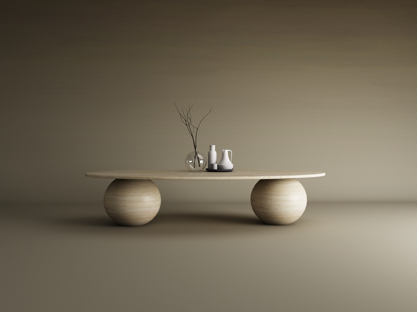 The TRAVERTABLE Ellipse Travertine Coffee Table with Stone Ball Base features a smooth, oval top and sculptural stone bases, styled with a glass vase of bare branches and two white ceramic pitchers on a black tray against a beige backdrop.