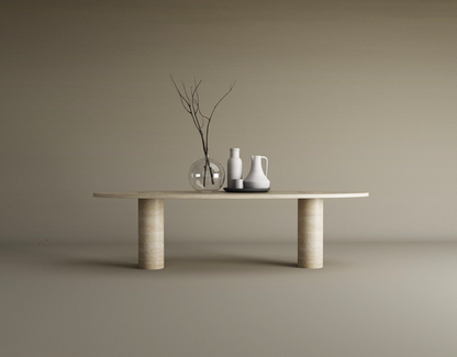 The TRAVERTABLE Ellipse Travertine Coffee Table with Solid Cylindrical Base showcases natural veining and supports a clear glass vase with branches, a white vase, and a white pitcher on a black dish, set against a plain beige background.