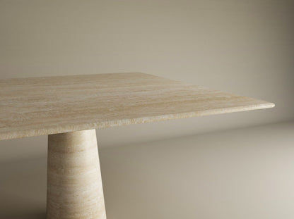 A close-up of the TRAVERTABLE Square Travertine Dining Table, featuring a natural travertine tabletop and a conical pedestal base, positioned against a plain light-colored wall. Customizable design.