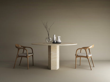 The TRAVERTABLE Oval Travertine Dining Table with a hexagonal center base brings timeless elegance, paired with wooden chairs and white cushions. Minimalist vases, like a clear vase with bare branches, complete the neutral beige-toned decor.