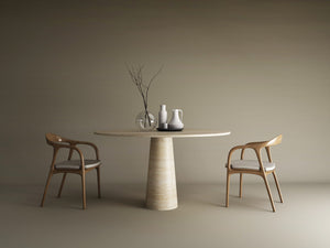 An oval natural travertine stone dining table by TRAVERTABLE with a conical base and two wooden chairs. On the tabletop: a clear vase with bare branches, a white jug, a gray jug, and a white cup, all against a beige background.