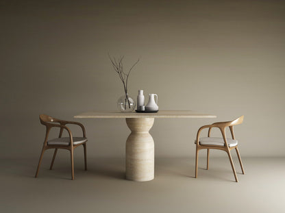 The Square Travertine Dining Table with Sculptural Round Center Base by TRAVERTABLE, featuring an organic stone form, is styled with vases and a pitcher. Two wooden chairs with white cushions flank this customizable table against a beige backdrop.