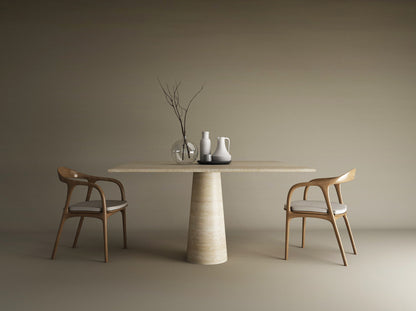 A minimalist scene featuring the TRAVERTABLE Square Travertine Dining Table with a conical pedestal base, natural travertine top, two wooden chairs with white cushions, and decorative vases and bare branches against a plain beige wall.