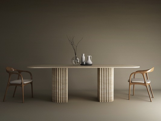 A minimalist dining scene features the TRAVERTABLE Rectangular Travertine Dining Table with Fluted Round Base, solid travertine stone base, two wooden chairs with light cushions, and simple vases with a branch centerpiece against a neutral backdrop.