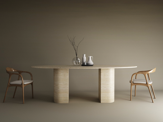A minimalist dining space highlights TRAVERTABLE’s Ellips Travertine Dining Table with a natural stone base and sloped pedestal, paired with two wooden chairs. The table holds ceramic vases, a small bowl, and a glass vase against a neutral backdrop.