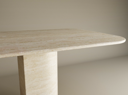 The TRAVERTABLE Rectangular Travertine Dining Table features a natural stone top with rounded corners and a thick sloped pedestal base, all crafted from solid travertine, standing out against a plain neutral background. Customization available.