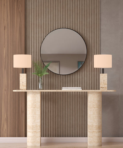 A TRAVERTABLE Rectangular Travertine Console Table with Sloped Pedestal Base, flanked by matching lamps and a glass vase of ferns, sits against a textured wall beneath a round mirror reflecting the room’s neutral decor.