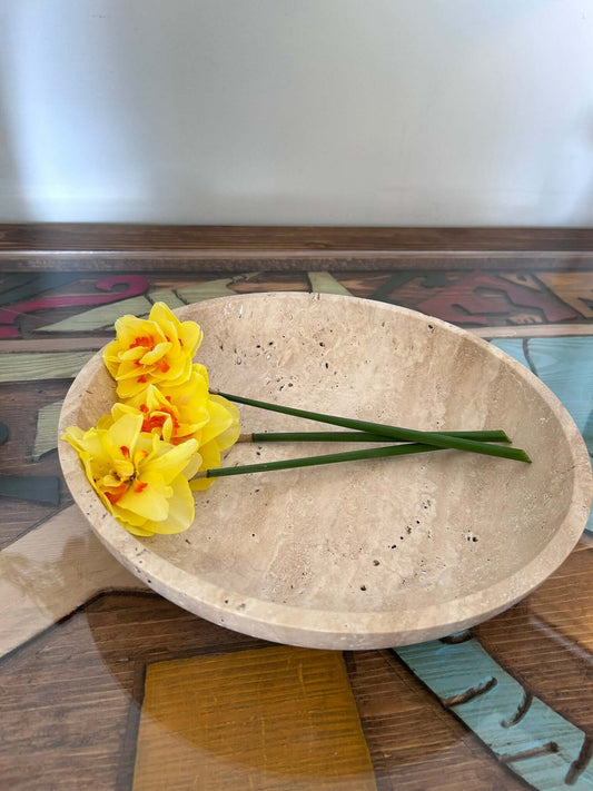 A Timeless Travertine Artisanal Bowl from Travertable sits on a colorful wooden table. Inside, three yellow daffodils with orange centers lie flat, showcasing natural artistry. The slightly blurred background highlights the bowl and flowers.