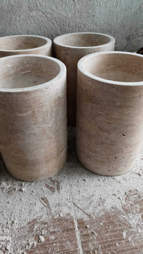 Four Golden Sunrise Travertine Vases by Travertable, featuring a smooth, light brown surface and subtle texture, stand on a dusty surface with a gray, uneven background.