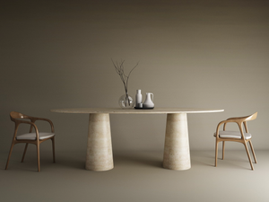 The TRAVERTABLE Ellips Travertine Dining Table with two conical stone bases stands amid wooden chairs with white cushions. Minimalist vases, bowls, and a glass branch vase sit atop the natural tabletop; the background is neutral.
