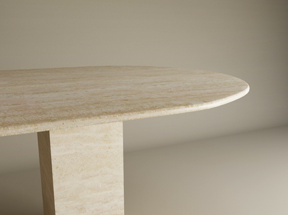 A close-up of the TRAVERTABLE Oval Travertine Dining Table, featuring a beige natural stone tabletop and a single rectangular travertine stone pedestal base, set against a plain, neutral background.
