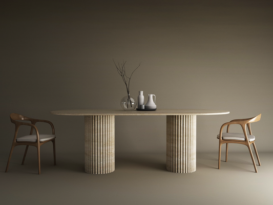 The TRAVERTABLE Oval Travertine Dining Table with Vertical Groove Base features a natural stone top, organic base, and is paired with two wooden chairs with white cushions. Minimalist decor includes a glass vase and ceramic pitchers against a beige background.