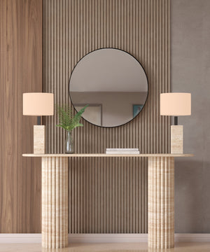 The Ovale Travertine Console Table by TRAVERTABLE features a vertical groove organic stone base and is customizable. Styled here with beige lamps, a glass vase with green leaves, and a round mirror on a wood slat accent wall behind it.