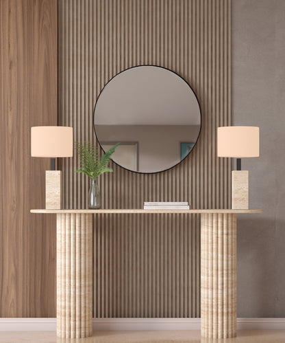 The Ovale Travertine Console Table by TRAVERTABLE features a vertical groove organic stone base and is customizable. Styled here with beige lamps, a glass vase with green leaves, and a round mirror on a wood slat accent wall behind it.
