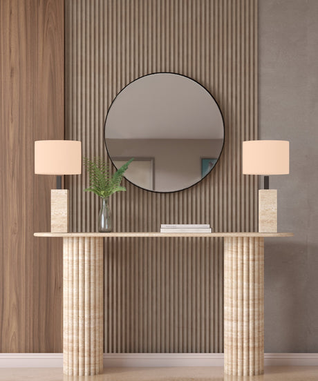 The Ovale Travertine Console Table by TRAVERTABLE features a vertical groove organic stone base and is customizable. Styled here with beige lamps, a glass vase with green leaves, and a round mirror on a wood slat accent wall behind it.