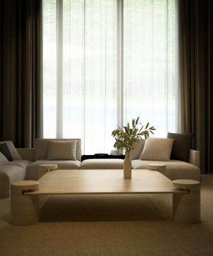 A modern living room features the TRAVERTABLE Square Travertine Coffee Table with Cylindrical Legs, crafted from pure travertine stone. Surrounded by beige sofas and neutral pillows, floor-to-ceiling windows with sheer curtains fill the space with soft light.