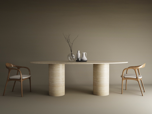 Minimalist dining setup showcasing the TRAVERTABLE Ellips Travertine Dining Table with a smooth cylinder base and pure travertine stone, paired with two wooden chairs with white cushions and decorative vases against a neutral beige background.