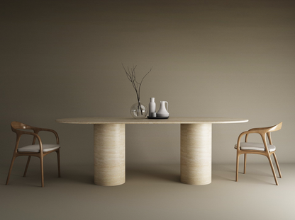 A minimalist dining setup featuring the TRAVERTABLE Oval Travertine Dining Table with Smooth Cylinder Base, two wooden chairs with white cushions, and simple vases—one holding bare branches—set against a plain taupe background.