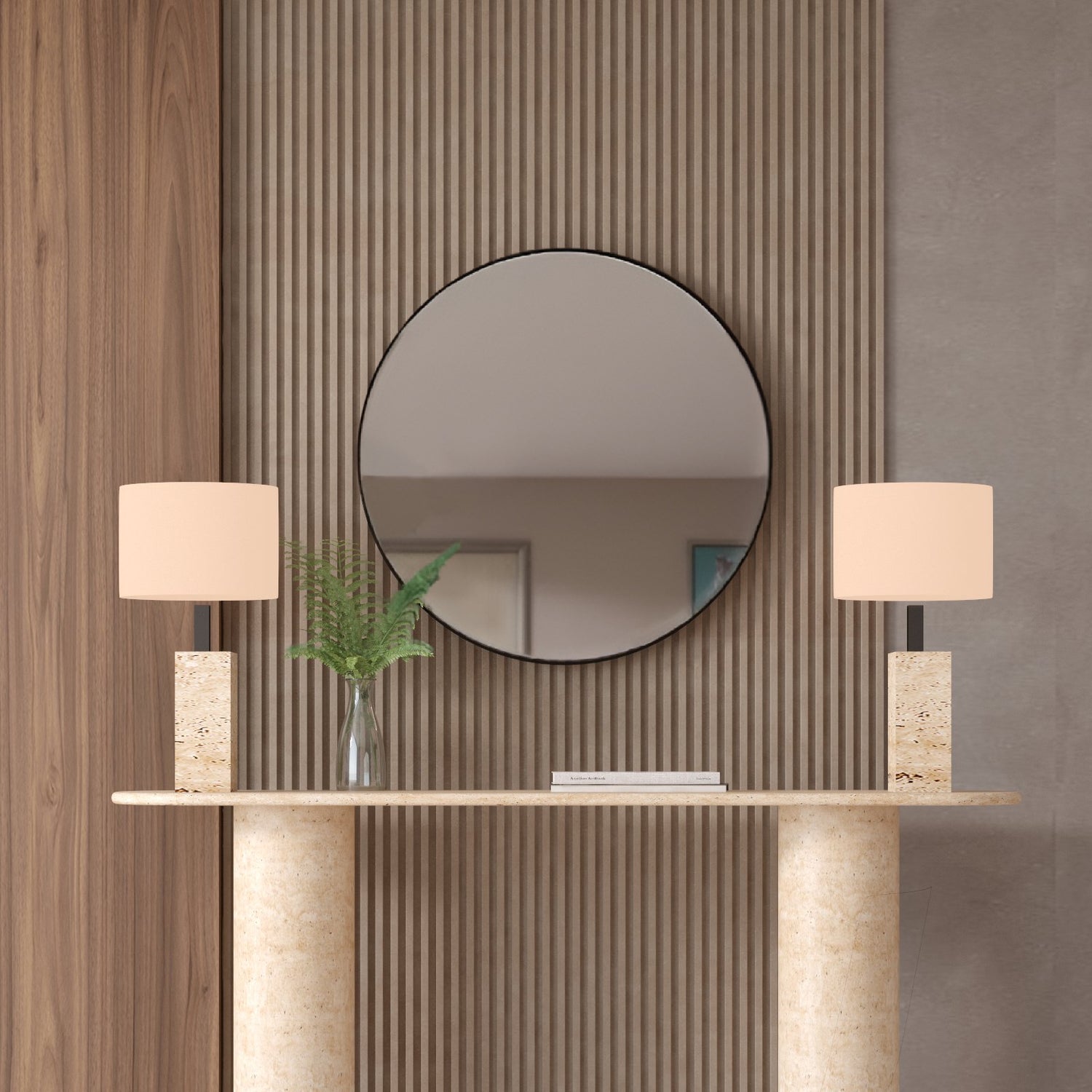 A round mirror hangs above the TRAVERTABLE Ovale Travertine Console Table with a smooth pure stone base, flanked by two stone lamps and a vase of green fern leaves against neutral vertical wood and textured panels.