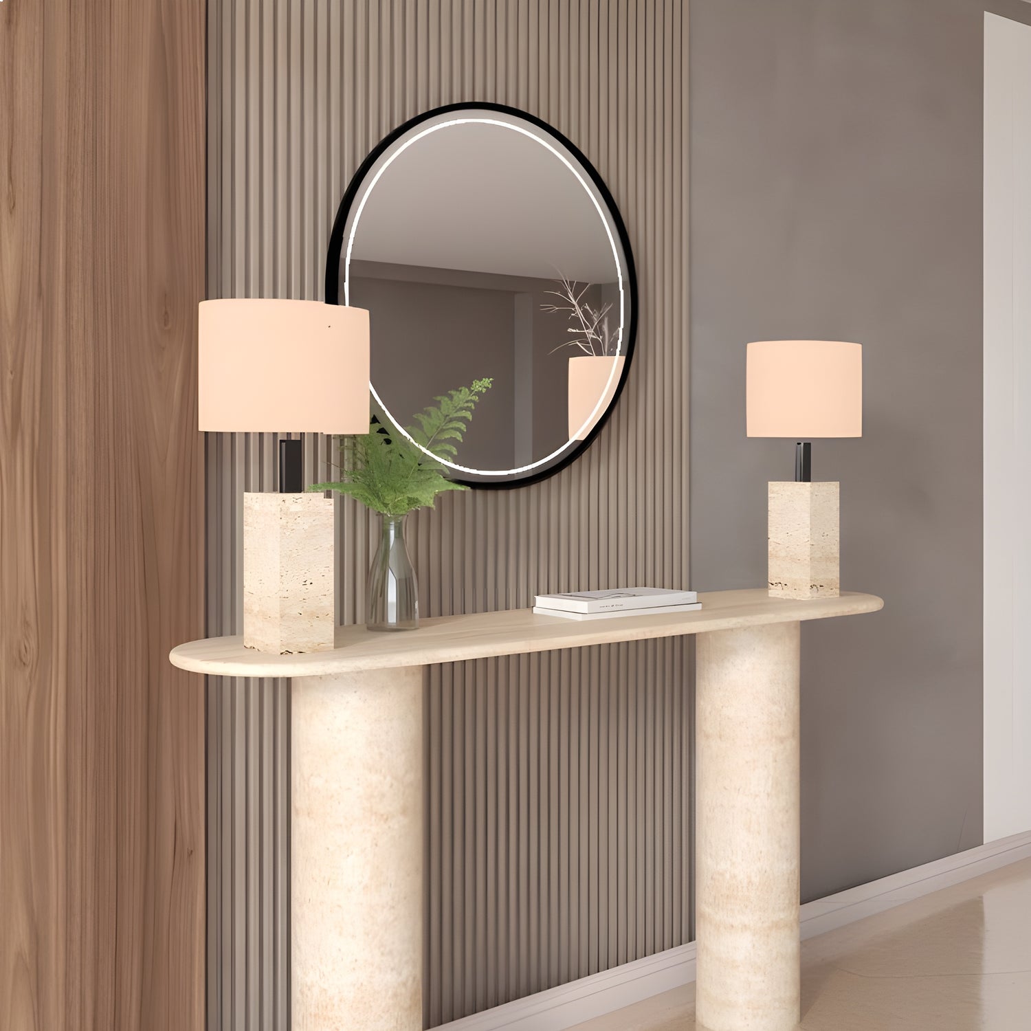 The TRAVERTABLE Ovale Travertine Console Table, with a pure travertine stone base and smooth cylinder design, holds two matching lamps and a glass vase of green foliage. Above, a round black-framed mirror reflects part of the room.